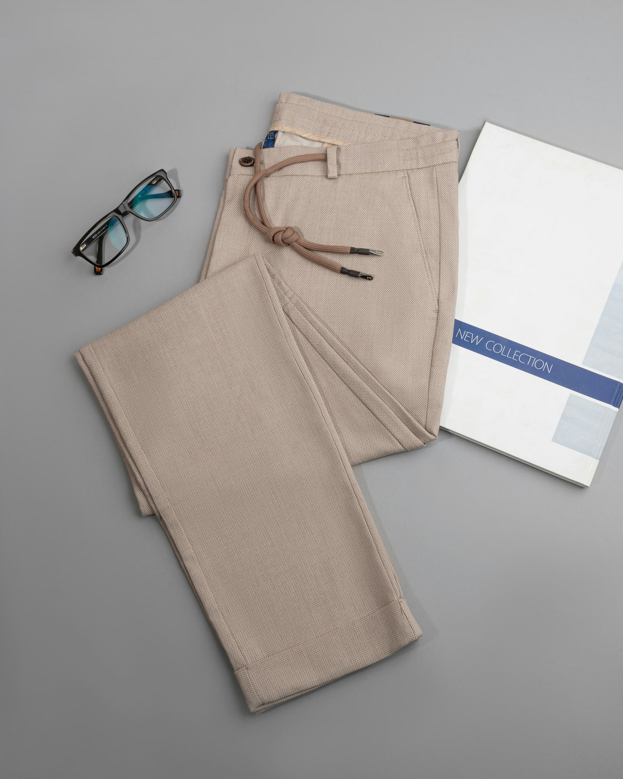 Stone drawstring trousers folded flat on grey beside glasses and a card