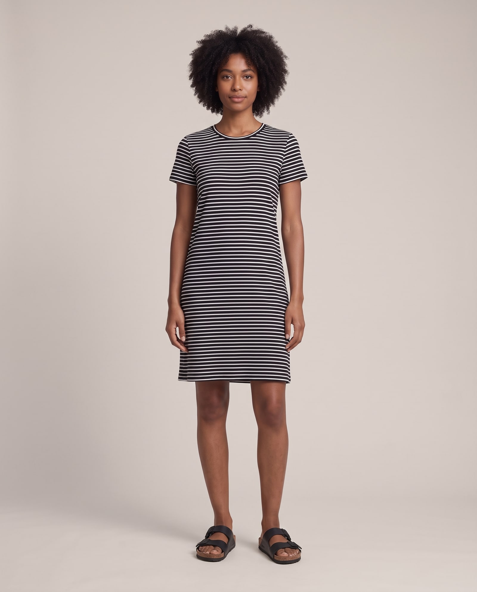 Model wearing the striped t-shirt dress with black sandals on a warm studio backdrop