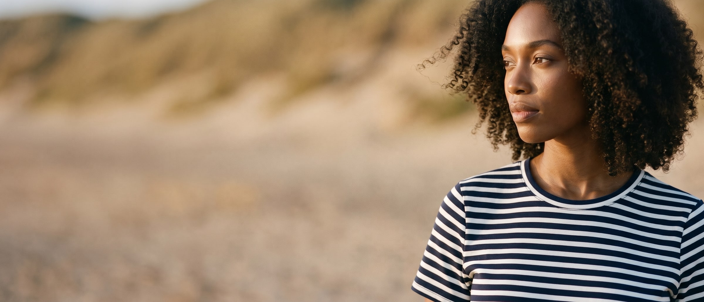 Close-up of a woman with curly hair in a navy and white striped t-shirt dress, her face and collar lit by low golden sun