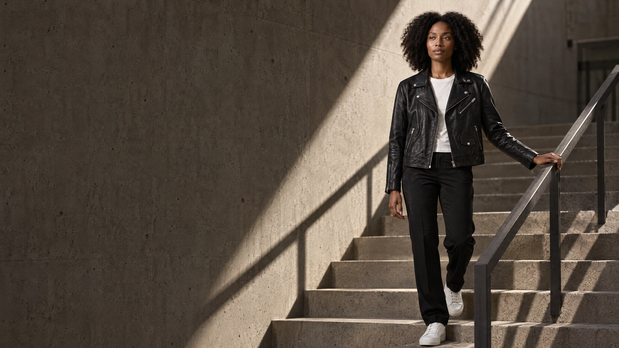 A woman with curly hair in a black leather biker jacket walking down a concrete stairwell through a shaft of hard light