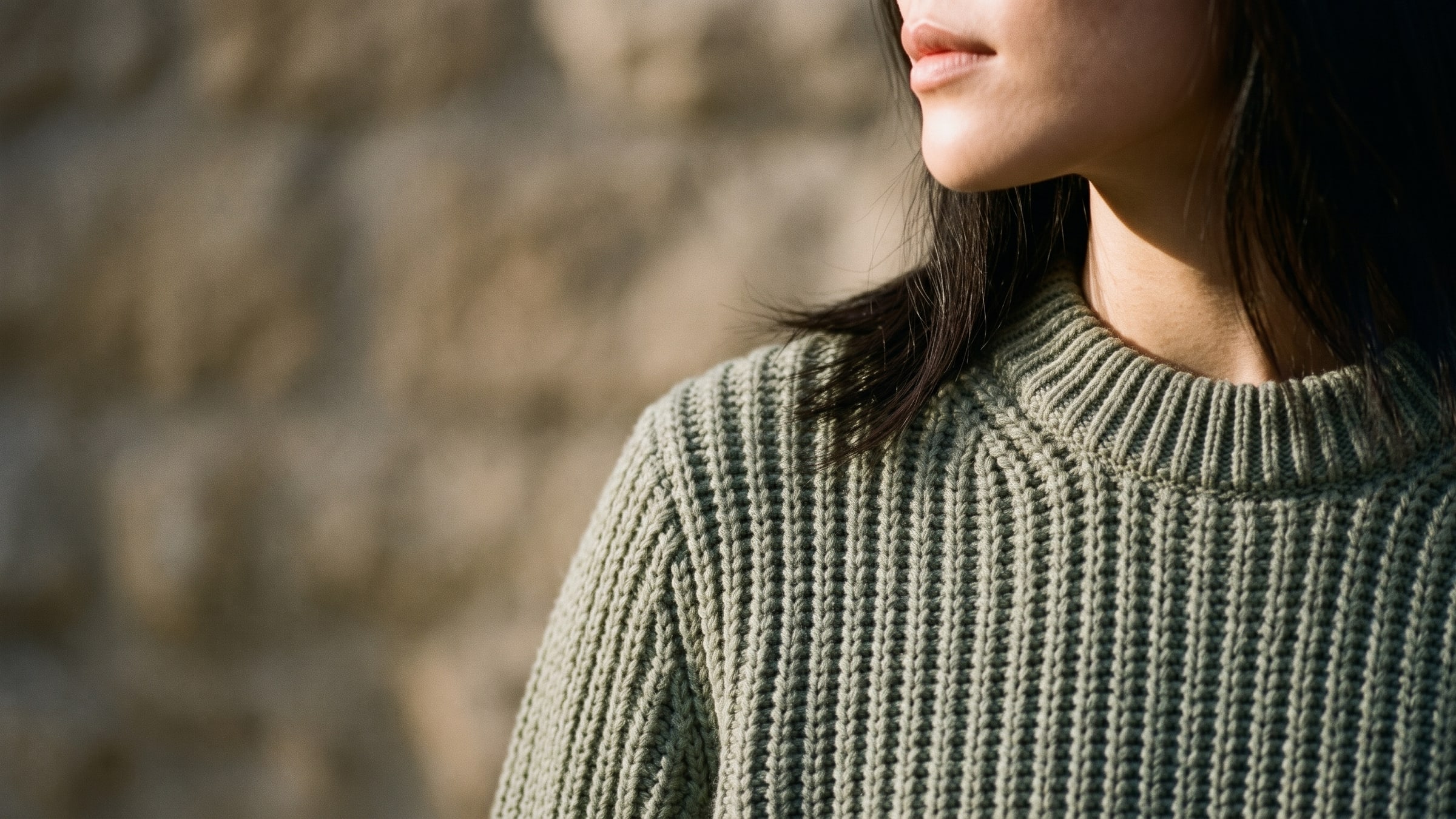 Close-up of a woman’s shoulder and collar in a sage ribbed crewneck, every rib of the knit lit by low raking sun