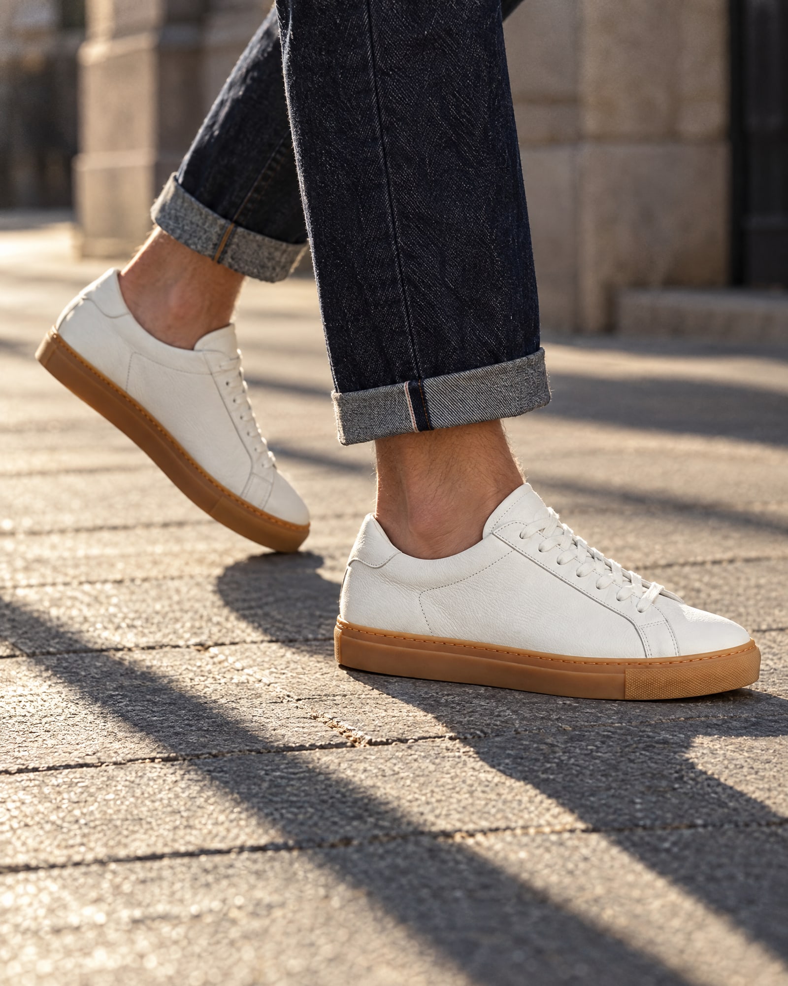 Ankle crop of white leather sneakers with gum soles and cuffed dark jeans mid-step on a sunlit pavement