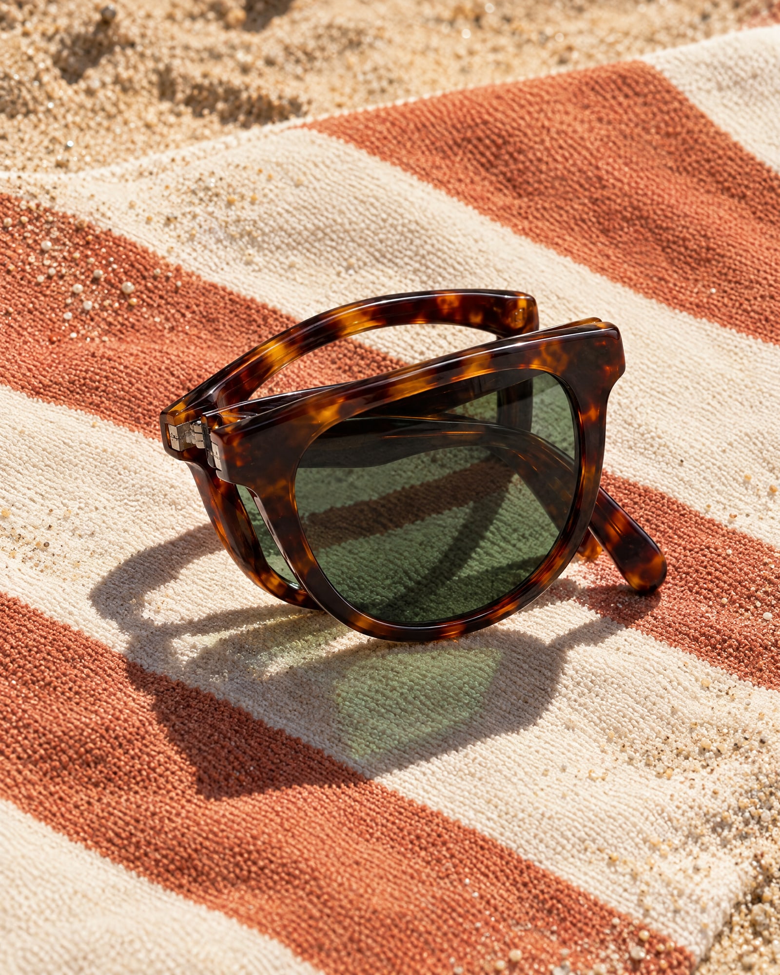 Folded tortoiseshell sunglasses with green lenses on a striped beach towel on the sand in hard sun