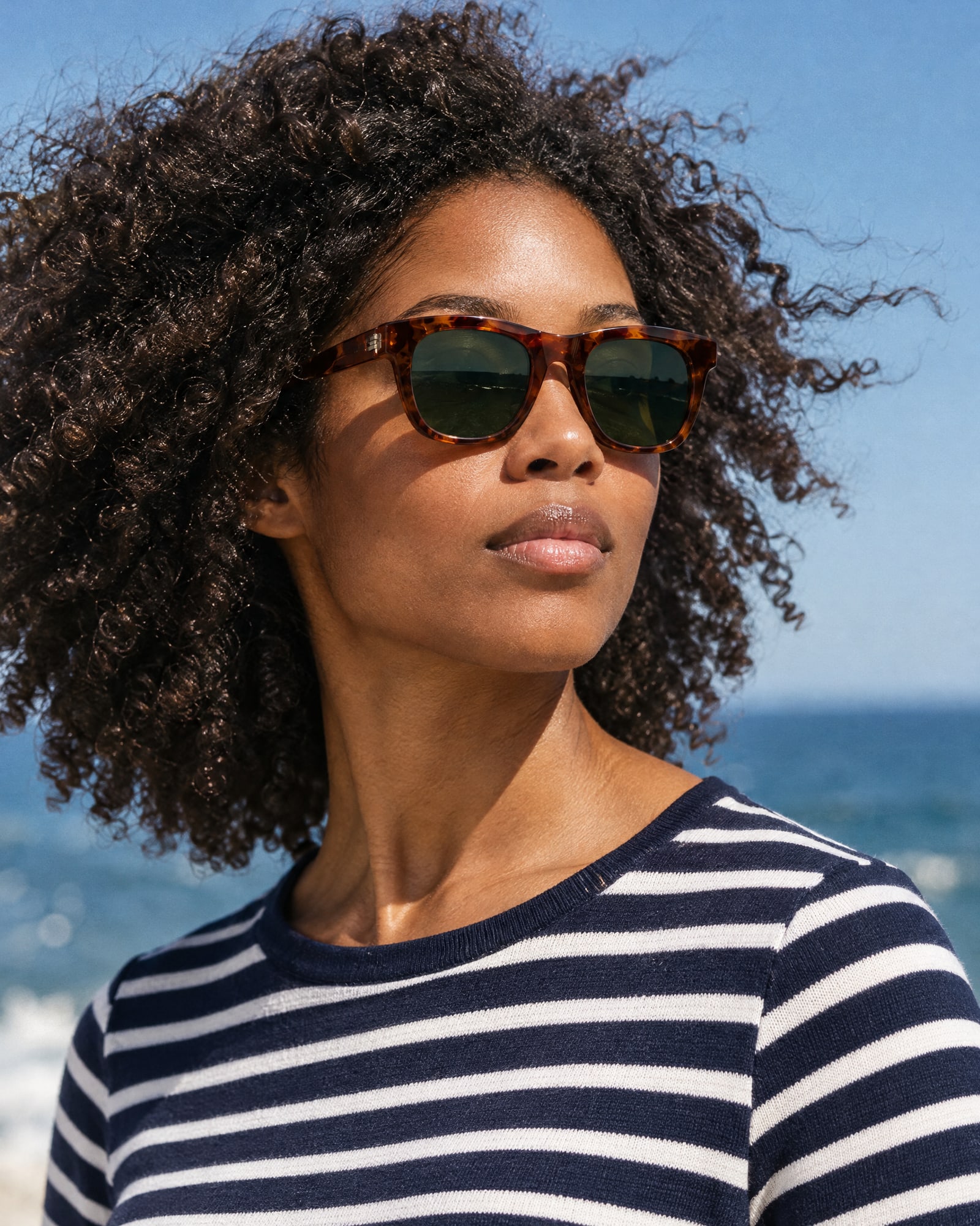 Close-up of a woman with curly hair wearing tortoiseshell sunglasses and a navy striped top by the sea, wind in her curls