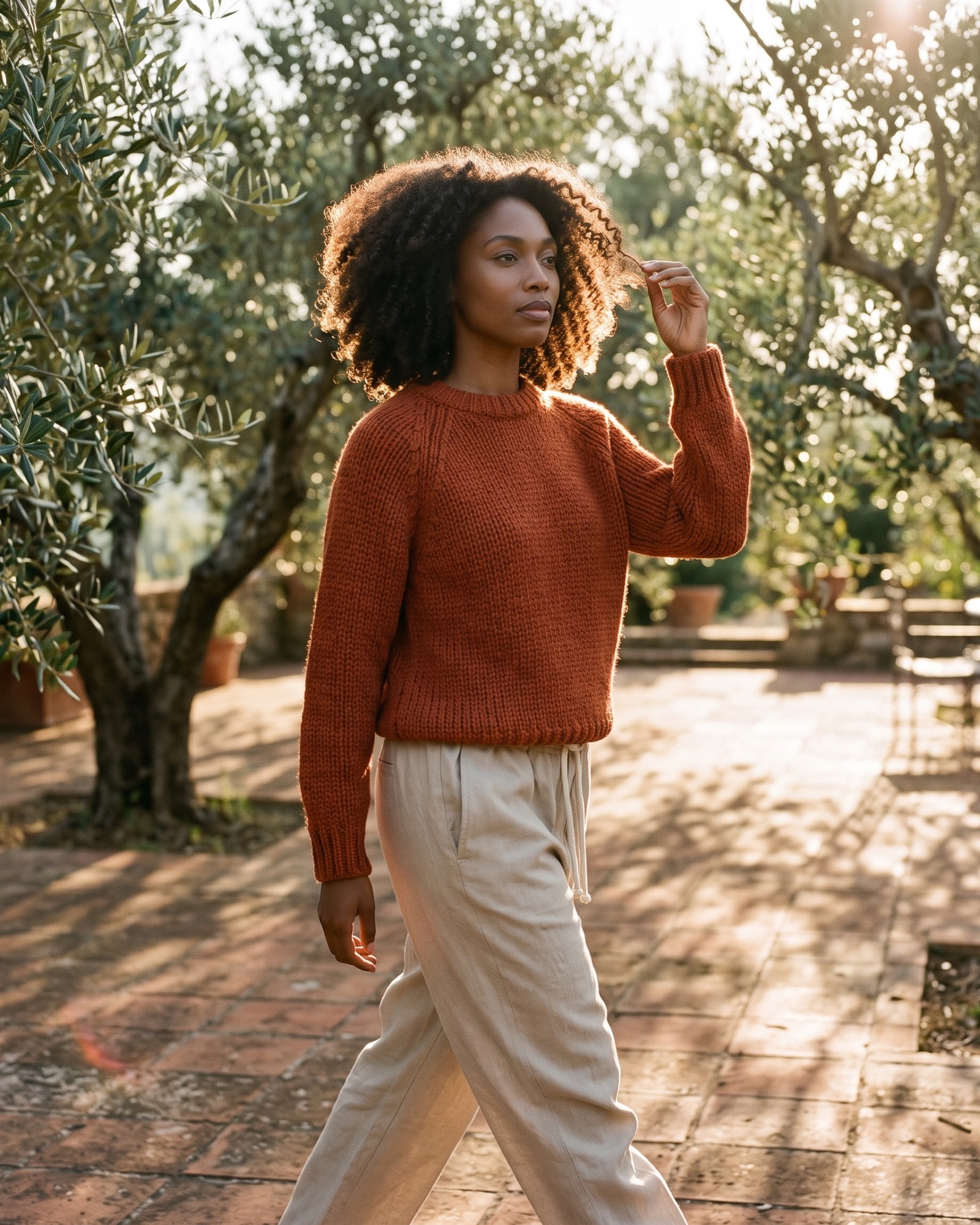 A woman with curly hair in a rust hand-knit sweater and stone drawstring trousers walking through an olive grove in golden evening light