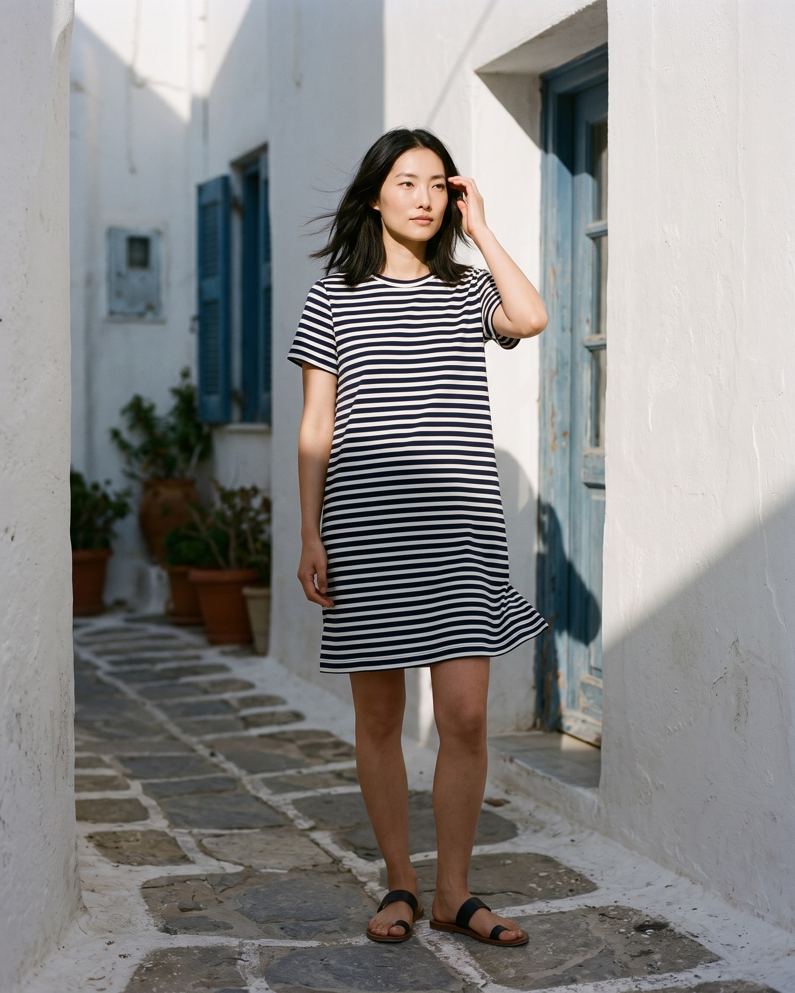 A woman in a navy and white striped t-shirt dress and sandals in a whitewashed Mediterranean alley with hard afternoon shadows