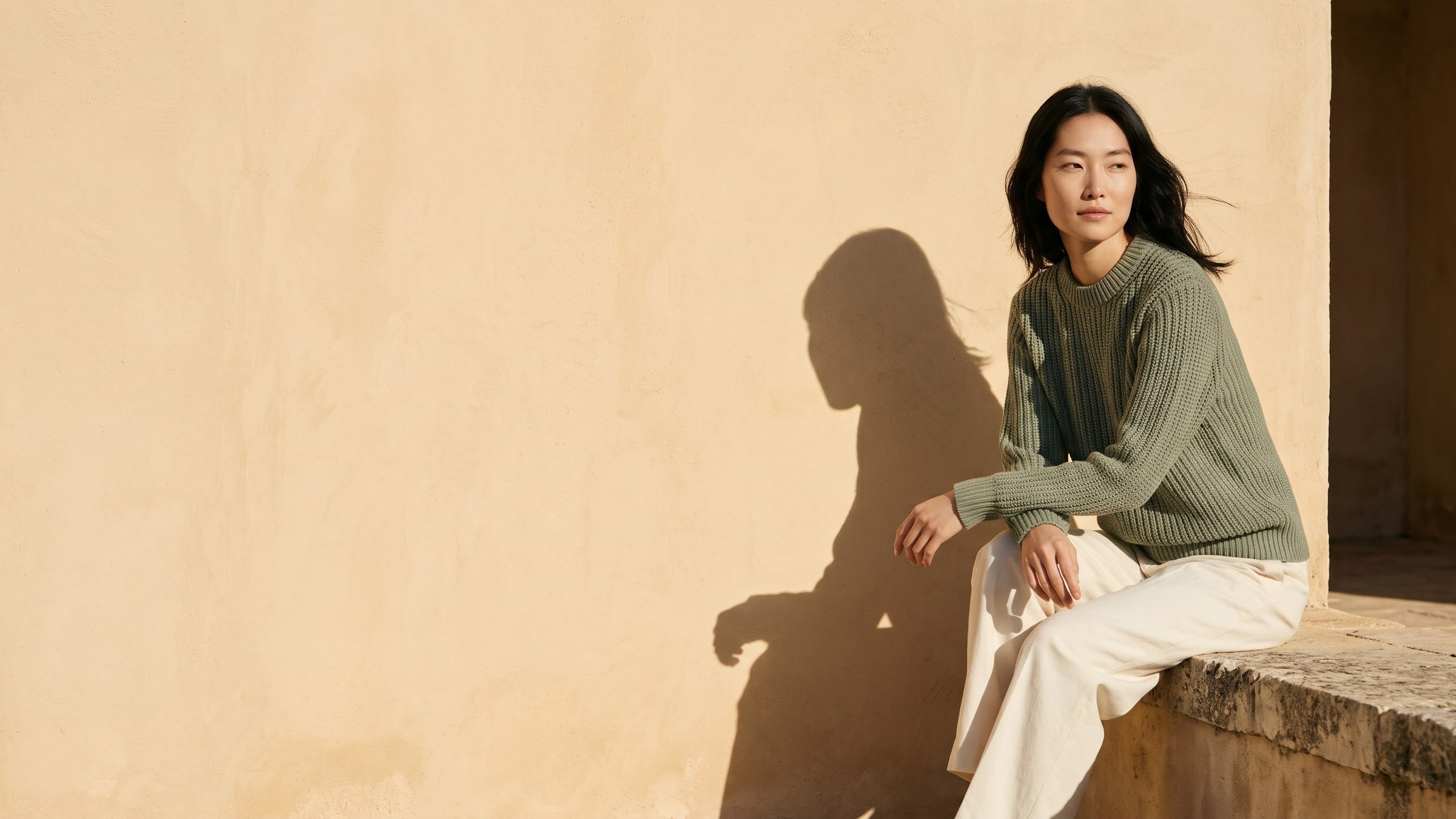 A woman in a sage ribbed crewneck and cream trousers sitting on a stone ledge against an ochre wall, her shadow cast beside her